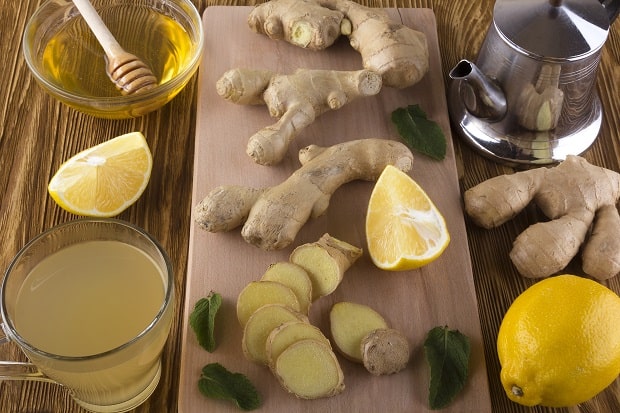кусочки свежего имбиря, лимон и листья мяты на разделочной доске, имбирный чай в стакане и заварник